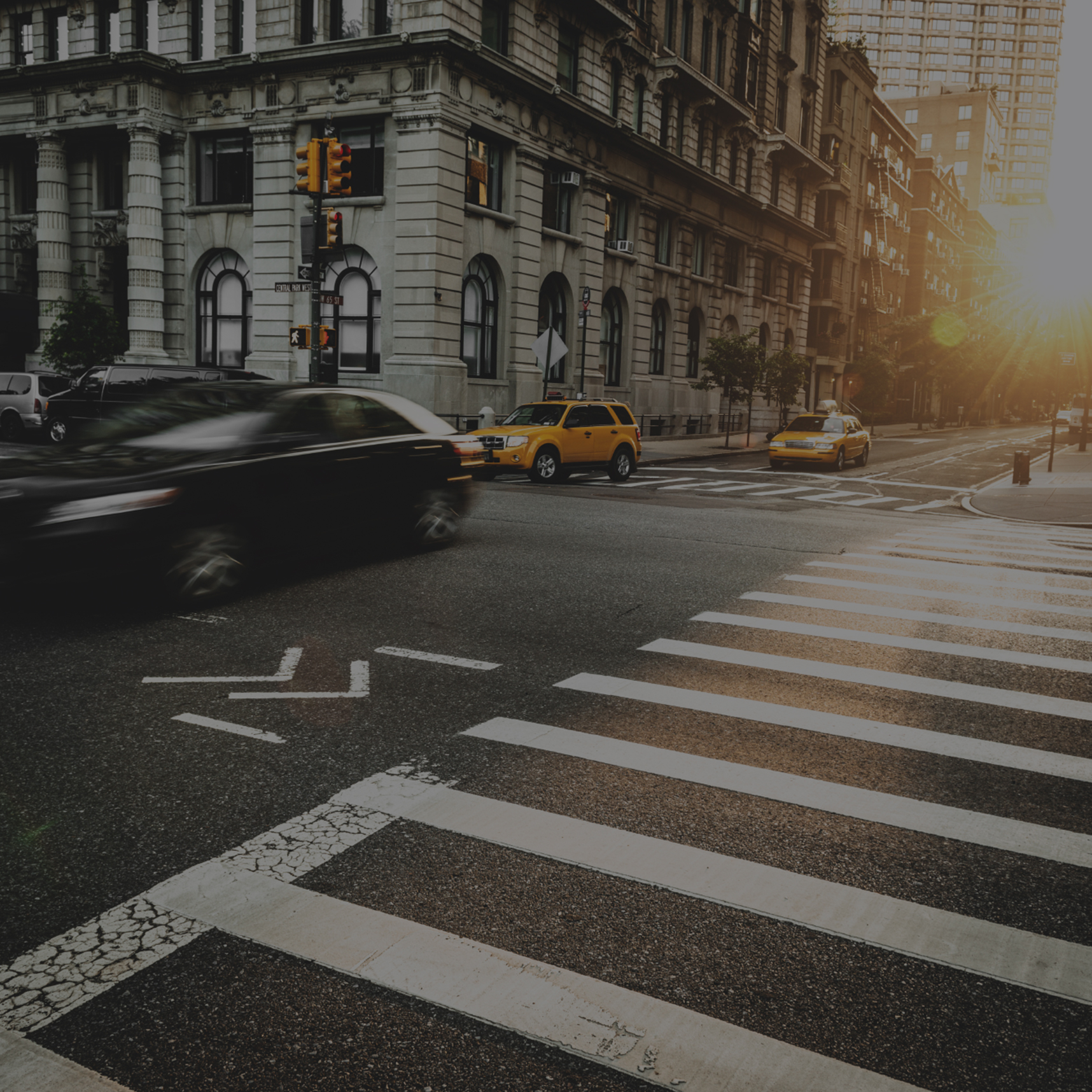 City street intersection at sunrise with blurred cars in motion, including a yellow taxi, and a pedestrian crosswalk in the foreground.